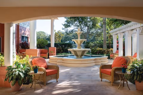 Outdoor seating area with wicker chairs featuring red cushions and small side tables with potted plants, surrounding a large tiered water fountain. The area is framed by columns and a pergola, with greenery and trees in the background under a clear sky.