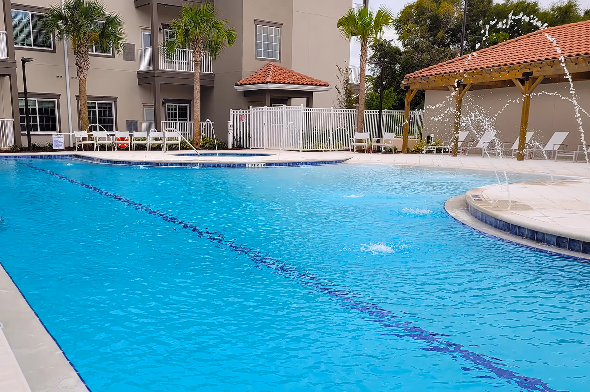 Outdoor swimming pool area with clear blue water, water fountains spraying arcs into the pool, palm trees, white lounge chairs, and a building with balconies and a red-tiled roof in the background.