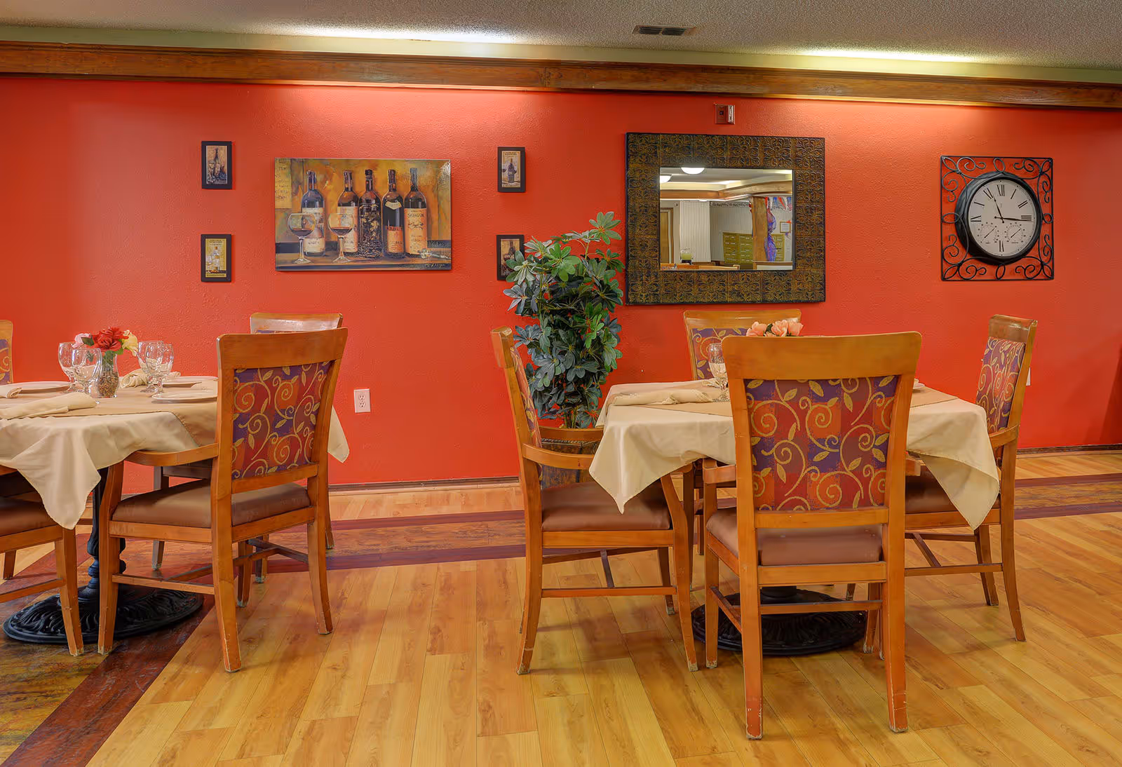 Dining area with tables set for meals, wooden chairs, and a red wall decorated with artwork, a mirror, and a clock.