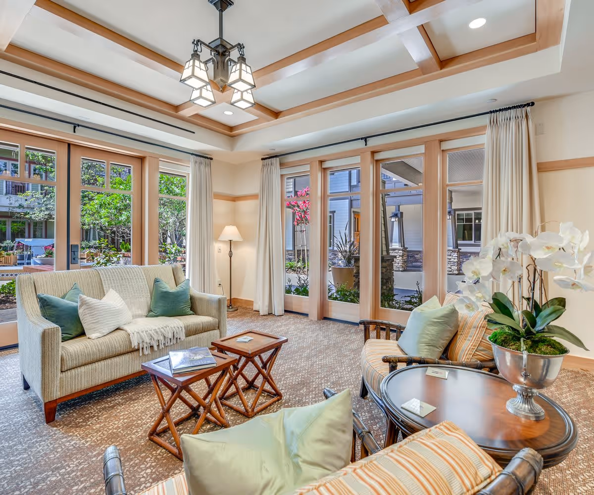 A bright and inviting living room with large windows and glass doors that open to a garden area. The room features a beige sofa with green and white pillows, two wooden nesting tables, a round wooden coffee table with a silver vase holding white orchids, and two cushioned armchairs with striped upholstery. The ceiling has wooden beams and a modern chandelier, and there is a floor lamp in the corner.