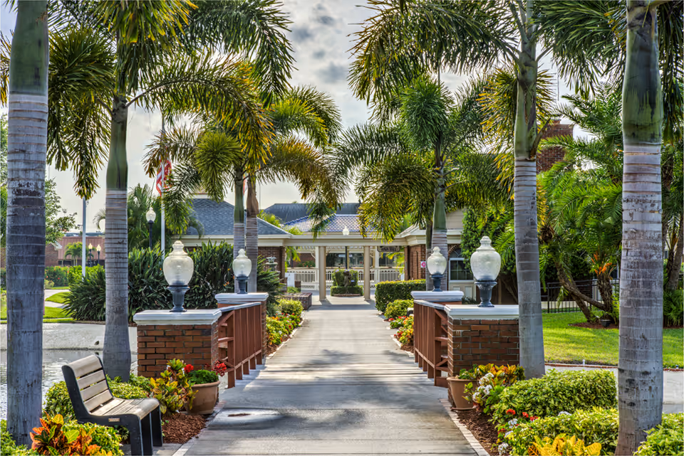 A paved walkway lined with palm trees and decorative lamp posts leads to the entrance of a building. There are benches and potted plants along the path, with well-maintained greenery and a small body of water on the left side. The sky is partly cloudy.