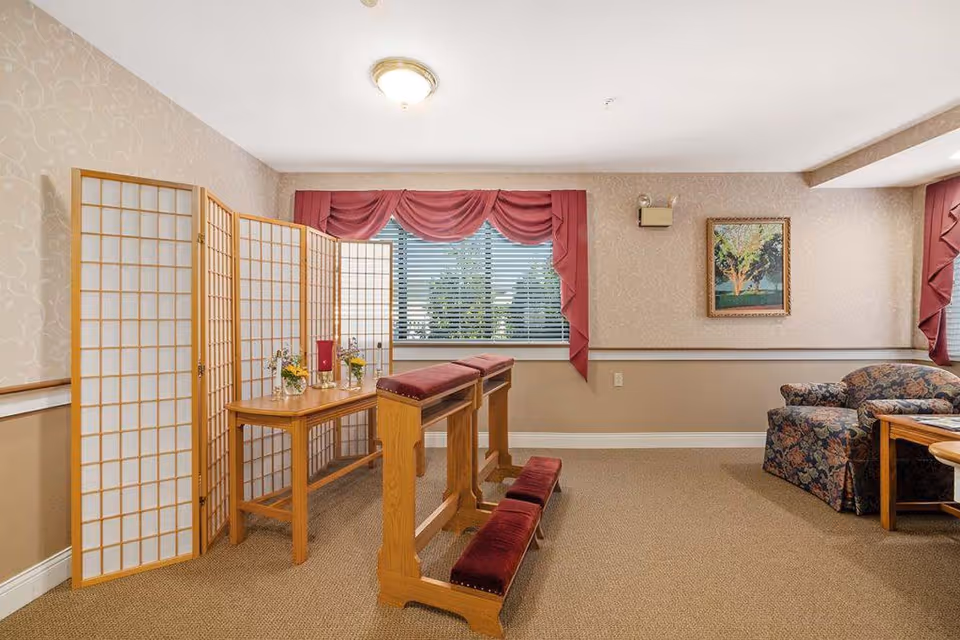 A quiet room with beige walls and carpet, featuring a wooden kneeling bench with red cushions, a wooden table with decorative items, a folding room divider, a window with red curtains, a floral armchair, and a framed painting on the wall.