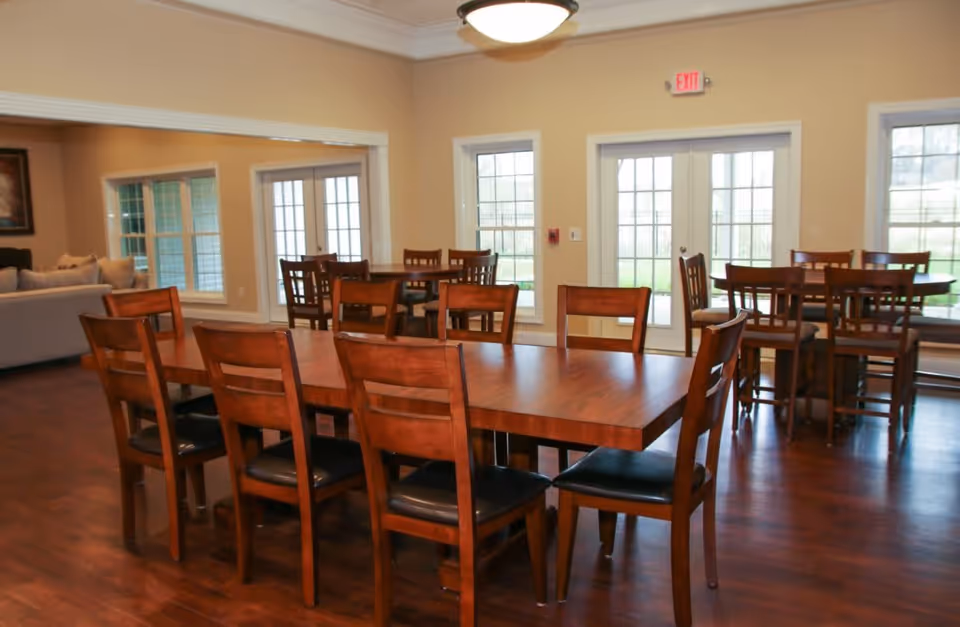 A dining area in Glenfield Memory Care featuring multiple wooden tables and chairs with dark cushioned seats. The room has large windows and glass doors allowing natural light to enter, with a beige wall color and wooden flooring. A seating area with a couch is visible in the adjacent room through an open wall space.