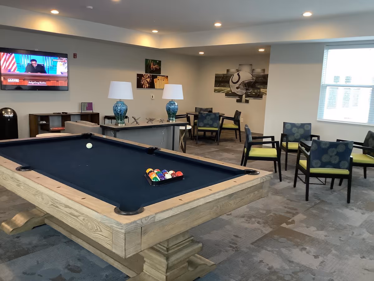 A recreational room with a pool table in the foreground, a flat-screen TV mounted on the wall, several chairs with green cushions arranged around small tables, two blue table lamps on a console table, and sports-themed wall art.