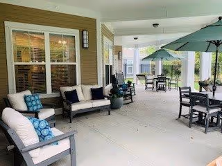 Covered outdoor patio area with cushioned seating including chairs and a loveseat, tables with umbrellas, and rocking chairs. The space is adjacent to a building with windows and a door, featuring light-colored flooring and walls.