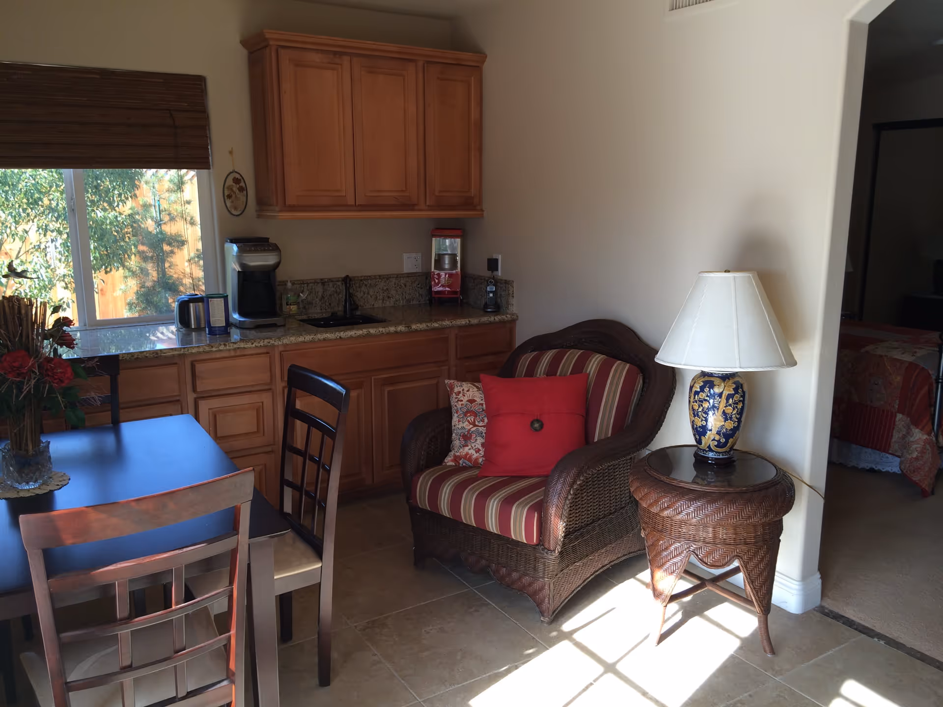 A cozy interior space featuring a small kitchenette with wooden cabinets, a granite countertop, a coffee maker, and a popcorn machine. In front of the kitchenette is a dark wooden dining table with chairs. To the right, there is a wicker armchair with red and striped cushions next to a wicker side table with a decorative lamp. A window with wooden blinds allows natural light to brighten the room. A bedroom with a bed covered in a red quilt is visible through an open doorway.