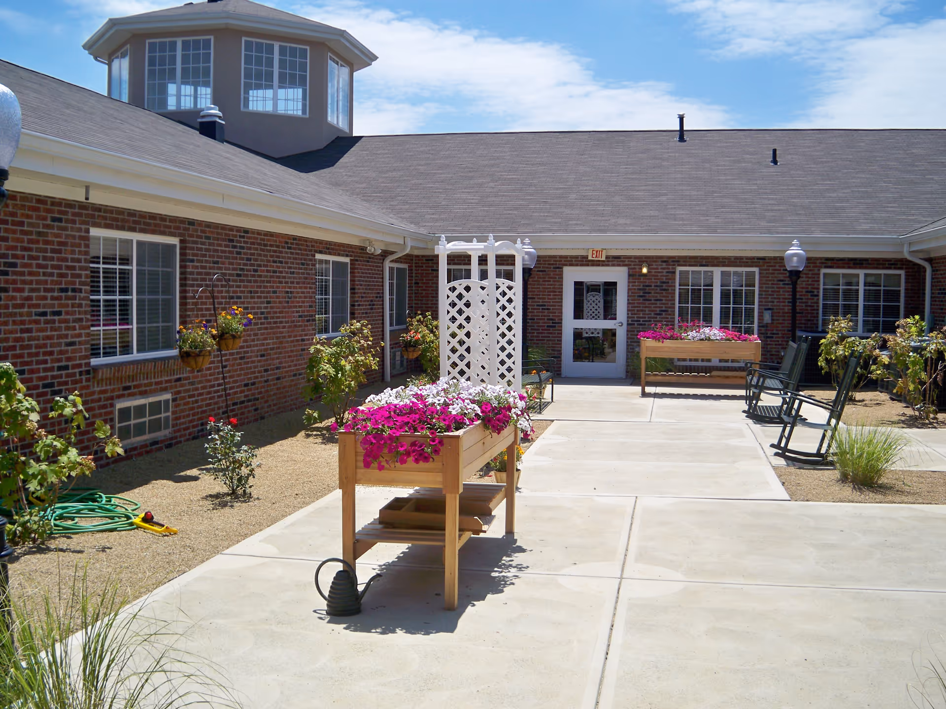 Outdoor courtyard area of a retirement community with brick walls, windows, and a door marked EXIT. The courtyard features flower beds with pink and white flowers, a white lattice trellis, green rocking chairs, and garden hose on the ground. The sky is clear with some clouds.