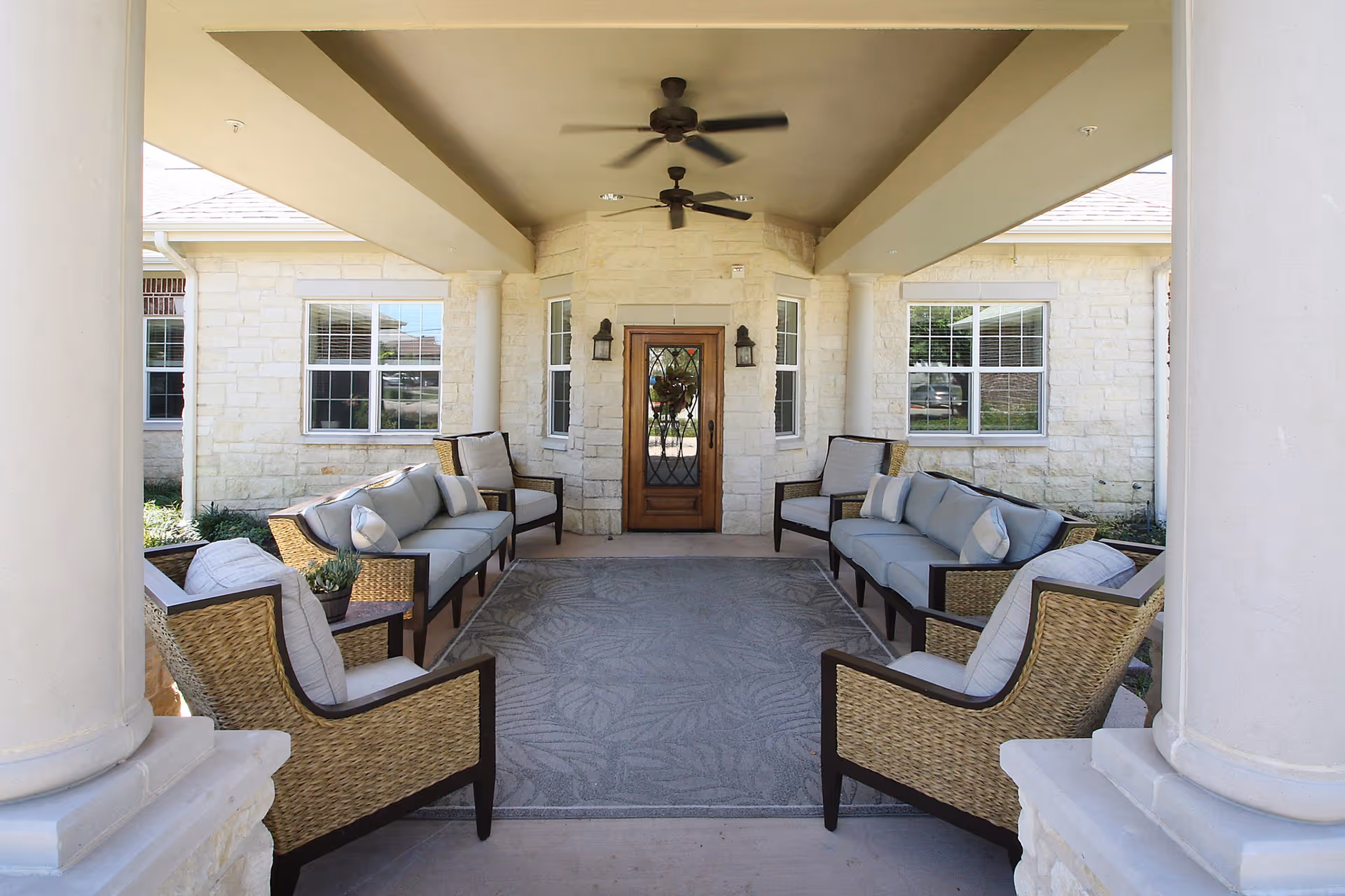Covered outdoor seating area with cushioned wicker chairs and sofas arranged around a large patterned rug, ceiling fans above, and a wooden door with glass panels at the far end, flanked by windows and stone walls.