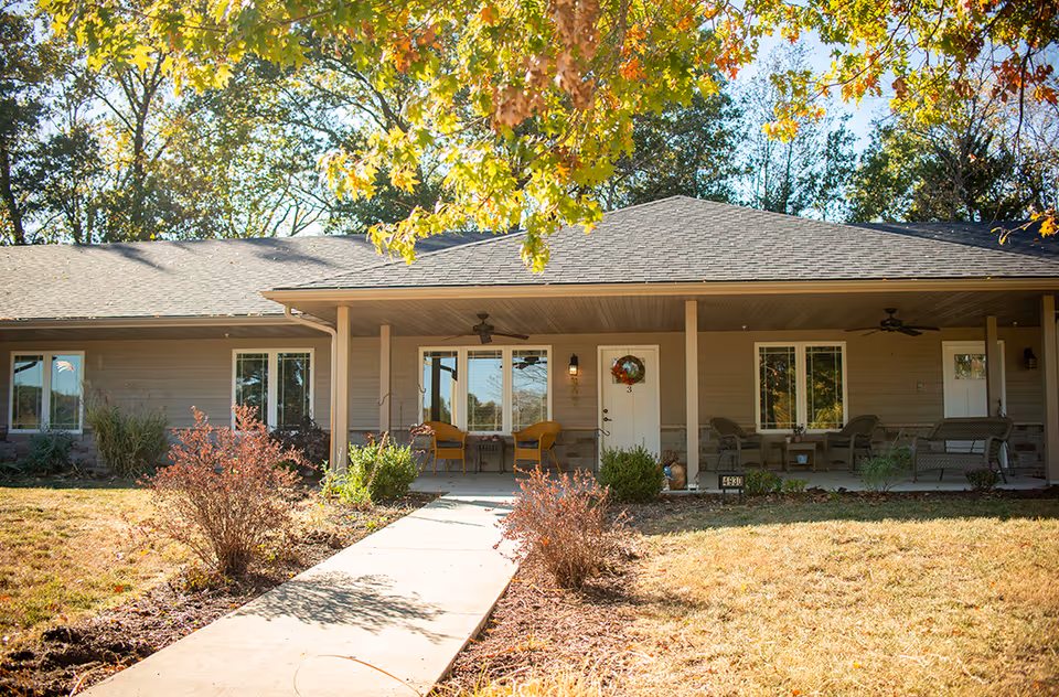 Single-story senior living building front with a covered porch, outdoor seating, a wreath-decorated door, and a walkway through a landscaped yard.