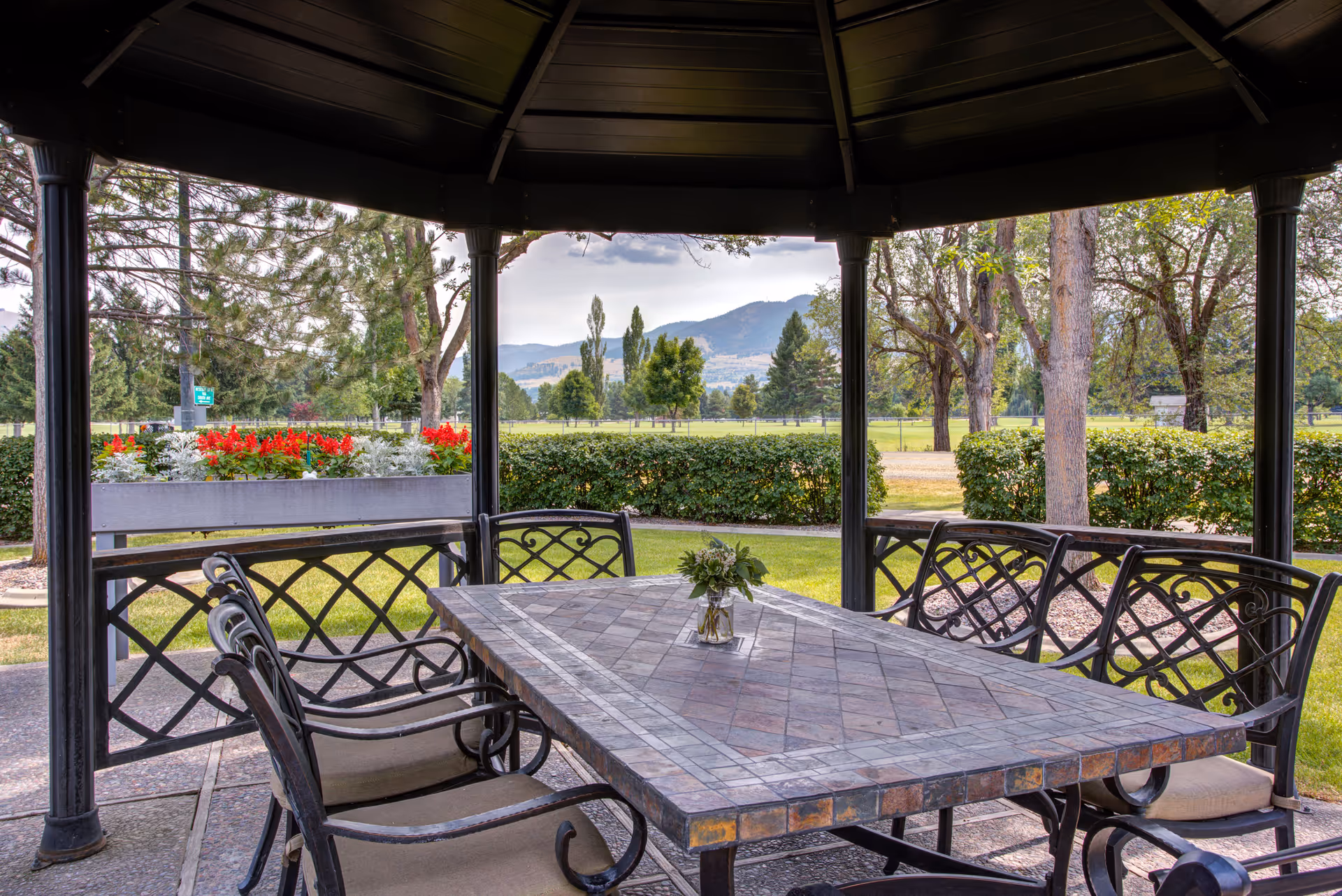 Outdoor gazebo with a large rectangular stone table surrounded by six metal chairs with cushions. A small vase with flowers is placed on the table. The gazebo overlooks a green lawn, bushes, trees, and distant mountains under a partly cloudy sky.