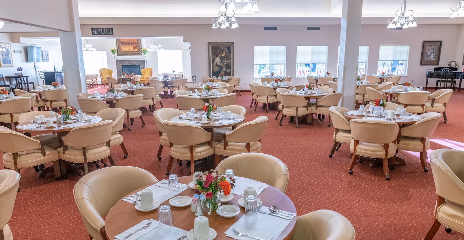 Large dining room with multiple round tables set for meals, beige chairs, red carpet, and floral centerpieces.