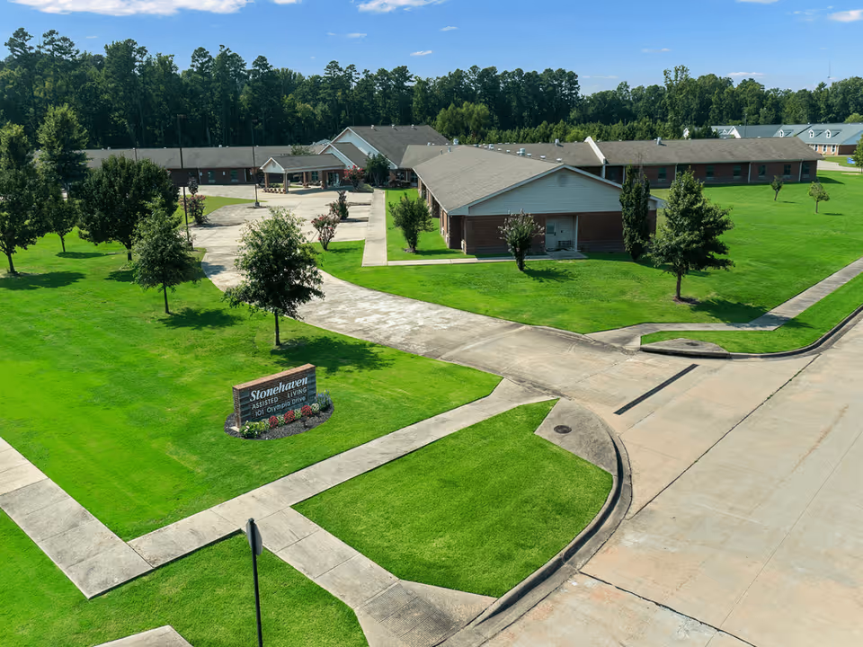 Aerial view of a senior living facility with multiple single-story brick buildings surrounded by well-maintained green lawns and trees. There is a paved driveway and sidewalks leading to the buildings. A sign in the grass reads 'Stonehaven Assisted Living 101 Olympic Drive.'