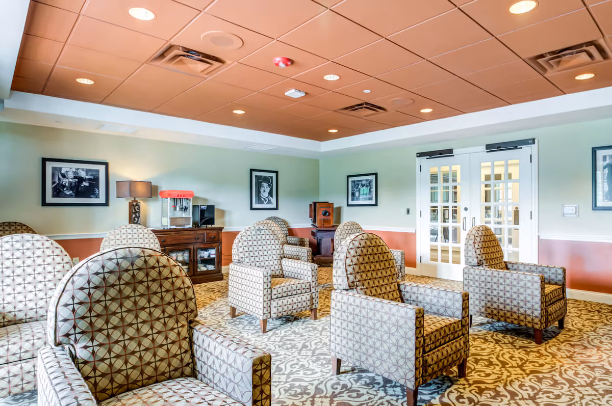 A bright senior living lounge with patterned armchairs arranged on a carpet, a sideboard with a popcorn machine, framed photos, and double glass doors.