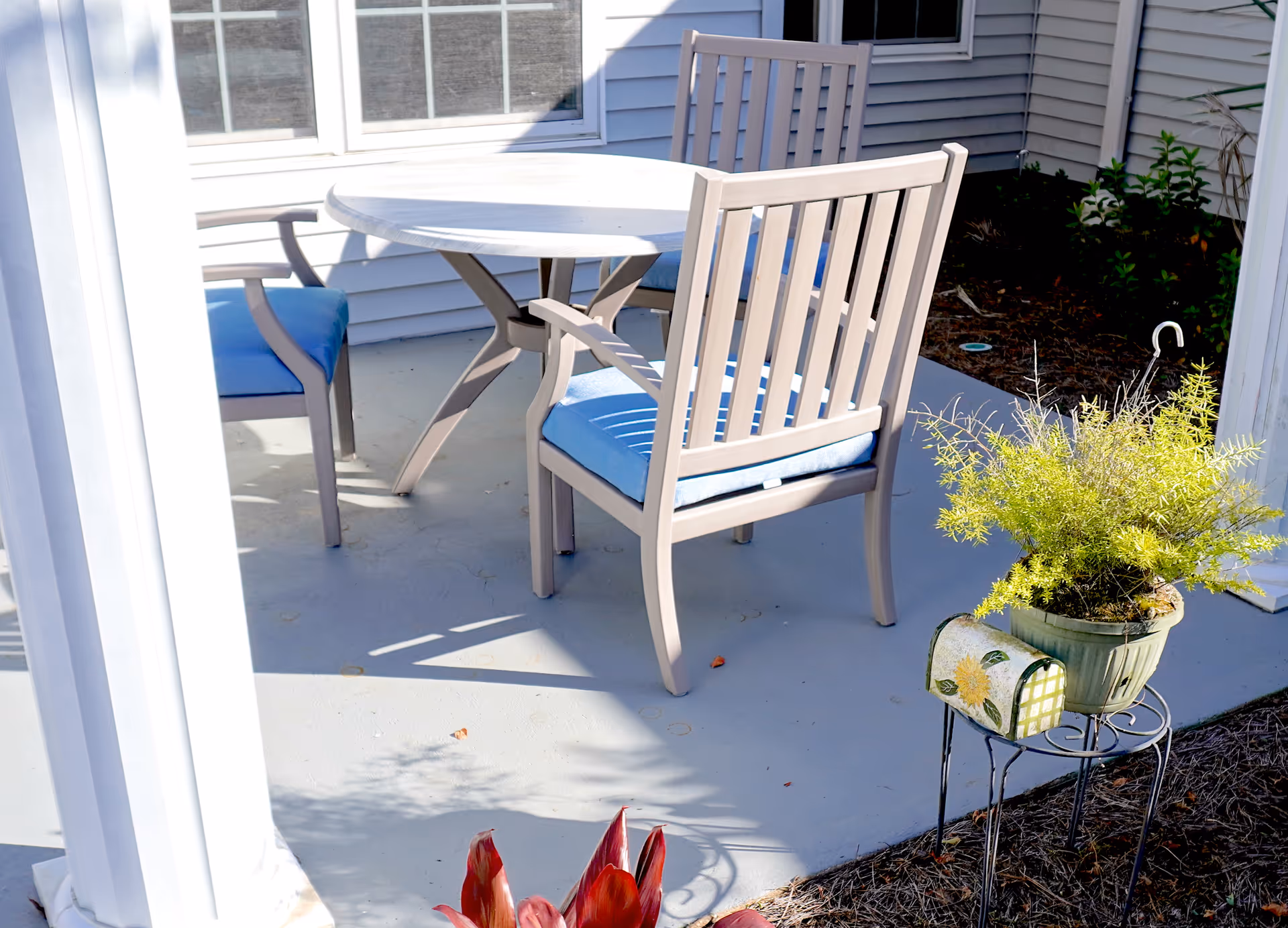 Outdoor patio area with a round white table and three beige chairs with blue cushions. The patio is adjacent to a white building with windows. There is a potted plant on a small metal stand and some garden plants nearby.