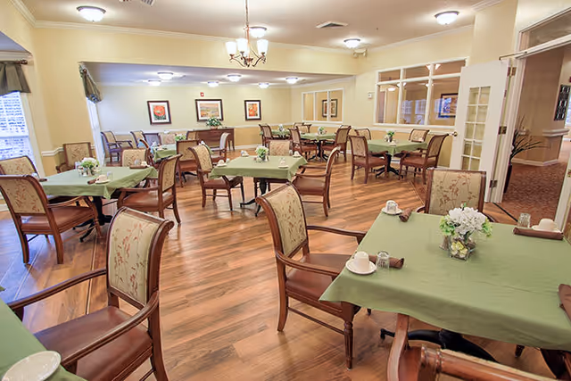 A spacious dining room with multiple tables covered in green tablecloths, each set with cups, napkins, and small flower arrangements. The room has wooden flooring, beige walls, framed artwork, and several chairs with floral upholstery. Ceiling lights and a chandelier provide warm lighting.