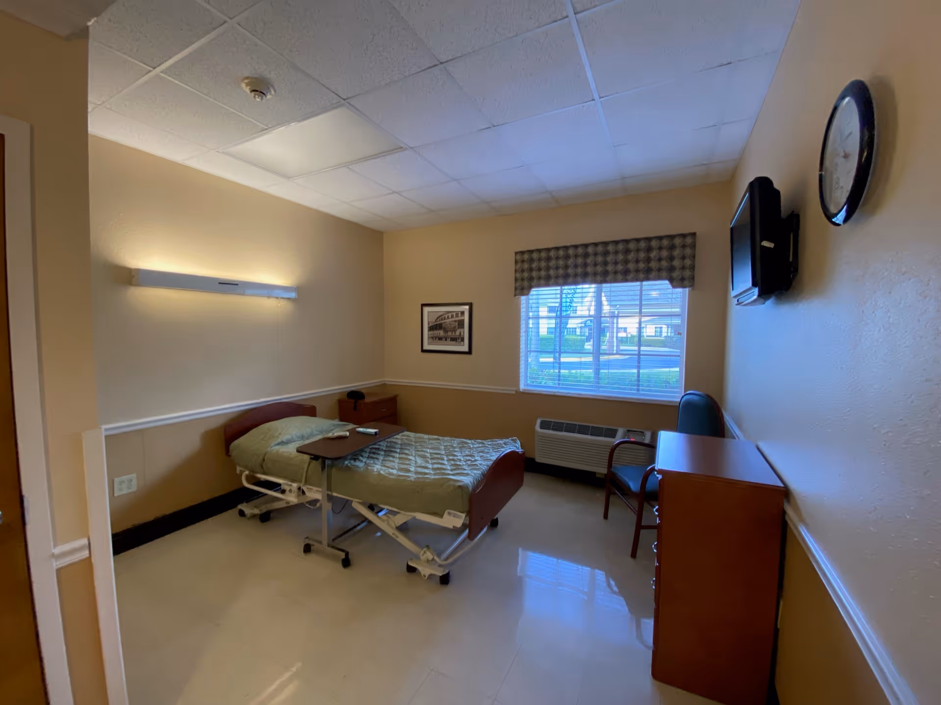 A small, simple bedroom in a senior living facility with a single hospital-style bed covered with a green quilt. The room has beige walls, a window with blinds and a valance, a wall-mounted TV, a clock, a wooden dresser, a chair, and a small bedside table. The floor is tiled and the ceiling has white panels.