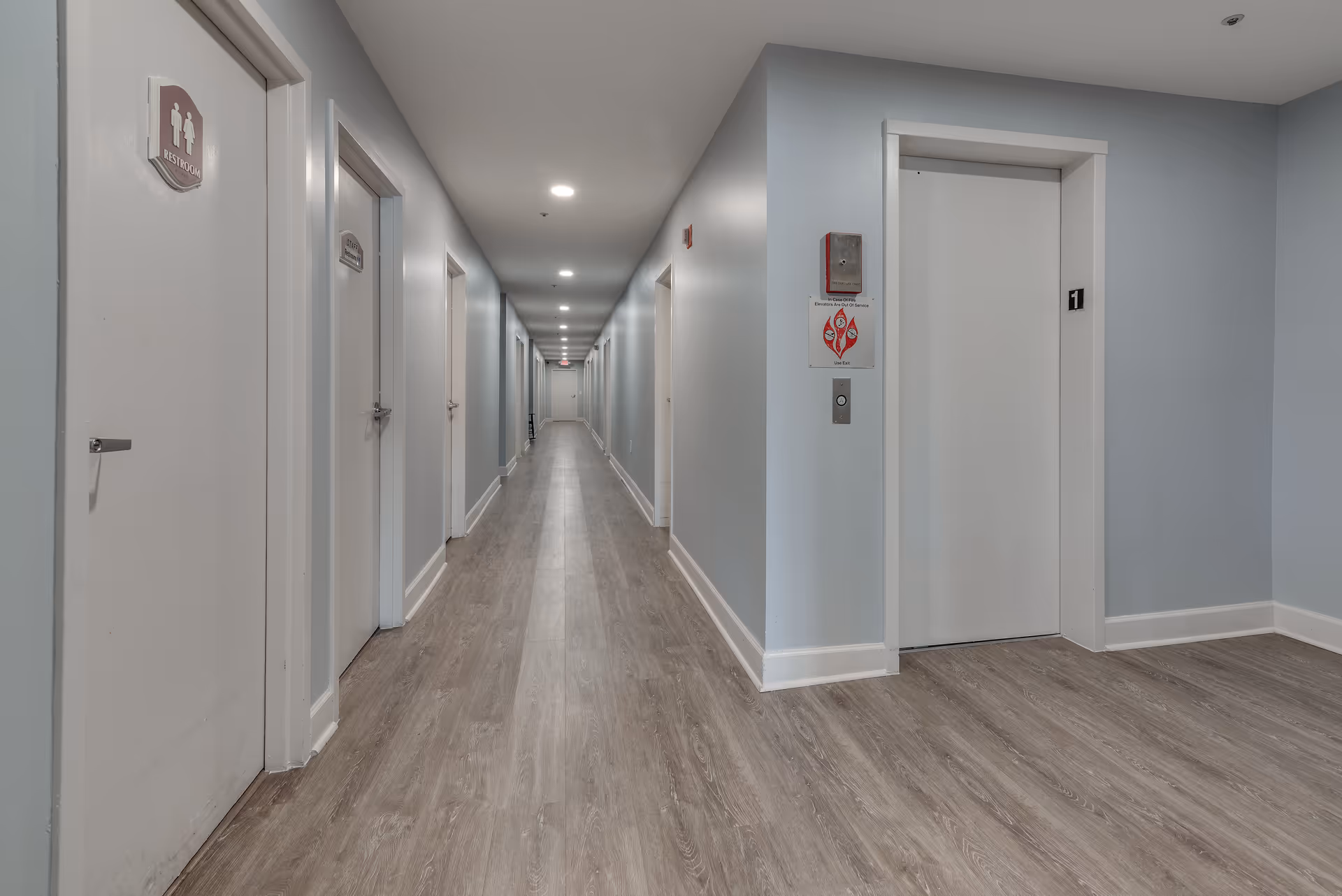 A long, well-lit hallway in a senior living facility with light blue walls and wood-patterned flooring. On the left side, there are several white doors including one marked as a restroom. On the right side, there is an elevator with the number 1 above it and a fire alarm panel next to it. The hallway extends into the distance with more doors visible along the walls.