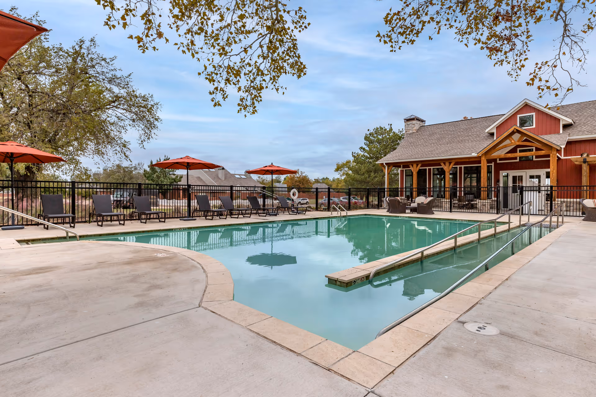 Outdoor swimming pool area with lounge chairs and red umbrellas around the pool. A building with a covered porch and wooden beams is visible in the background. Trees and a partly cloudy sky are also seen.