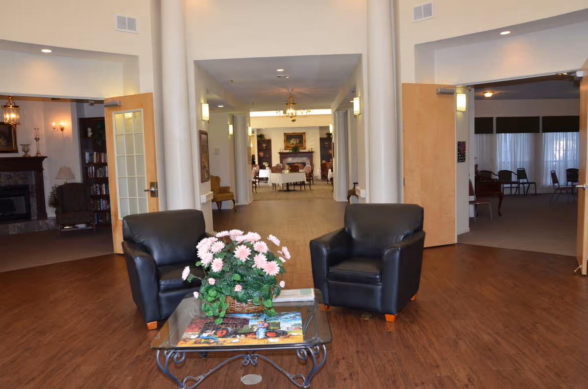 A spacious interior common area with two black leather armchairs facing a glass coffee table with a basket of pink flowers. The room has wooden flooring and leads into a hallway with tables and chairs, and a fireplace visible in the background. Open doors on either side reveal additional seating areas with chairs and lamps.