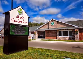 A sign reading 'Shields Comfort Care' stands on the lawn in front of a single-story brick assisted living building under a blue sky.