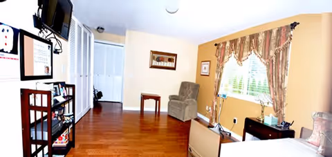 A well-lit interior room with wooden flooring, a recliner chair, a small wooden table, a window with floral curtains, a TV mounted on the wall, and a bed partially visible on the right side. The walls are painted in light beige and mustard colors.