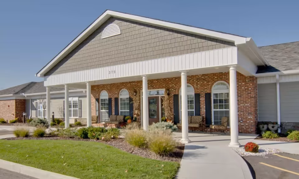 Front exterior view of Parkway Gardens Senior Living facility featuring a brick and siding facade, white columns supporting a covered entrance, landscaped greenery, and outdoor seating on the porch.