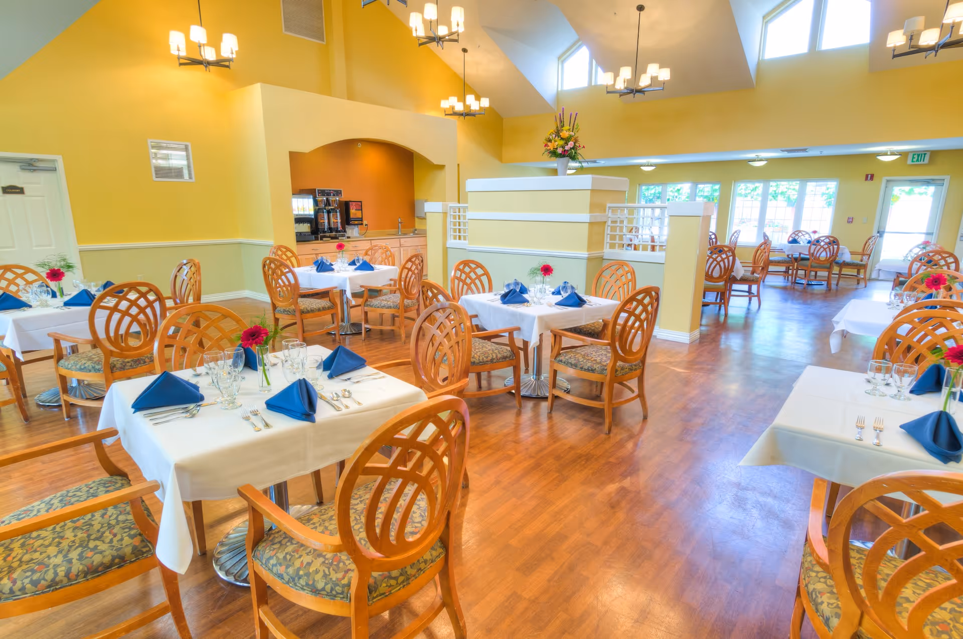 Bright and spacious dining room with wooden chairs and tables covered with white tablecloths, each set with blue napkins, glassware, and silverware. The room features yellow walls, large windows letting in natural light, and ceiling chandeliers. There is a coffee station in the corner and a floral arrangement on a partition.