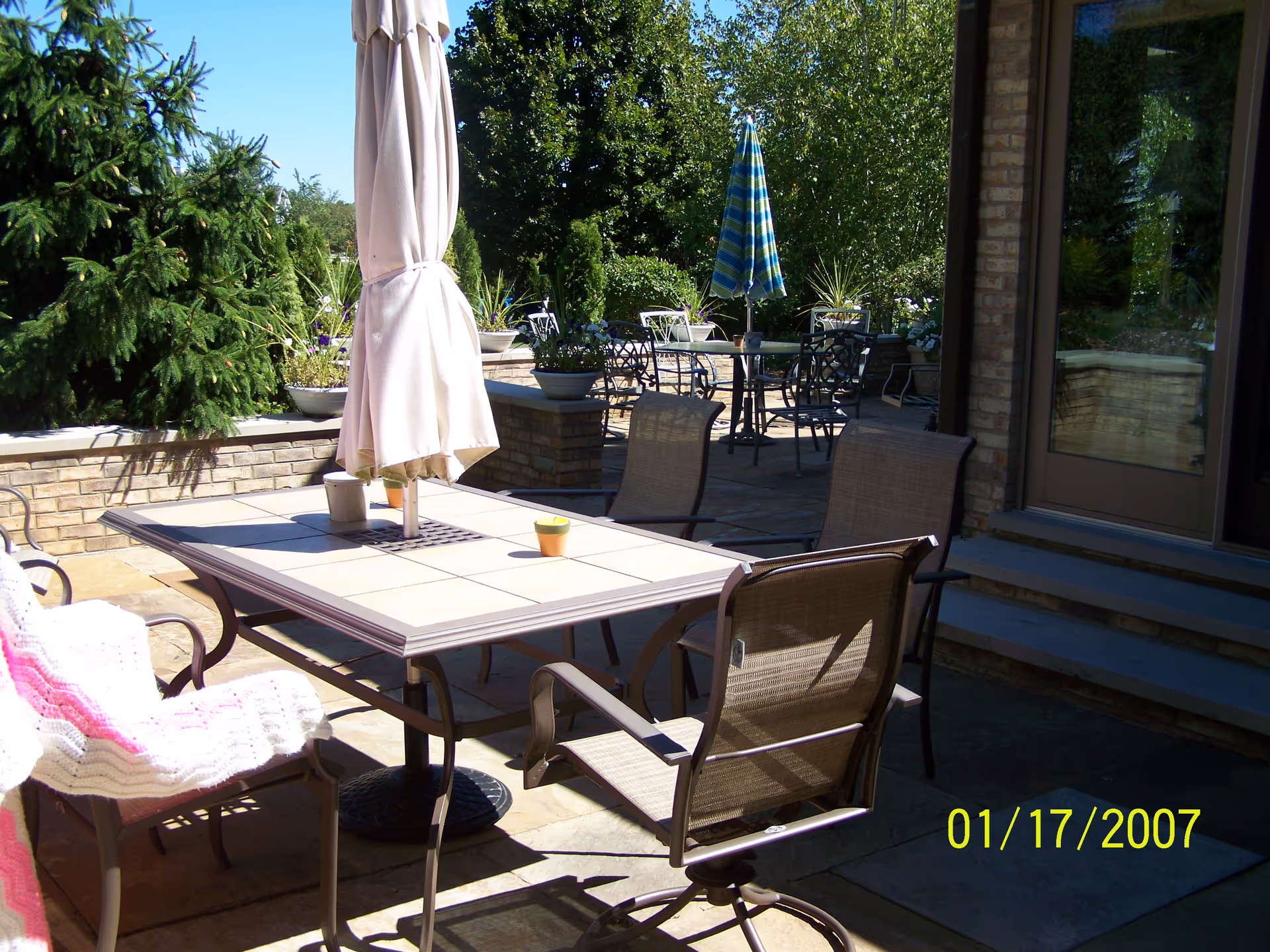 Outdoor patio with metal tables, chairs, umbrellas, and potted plants beside a brick building.