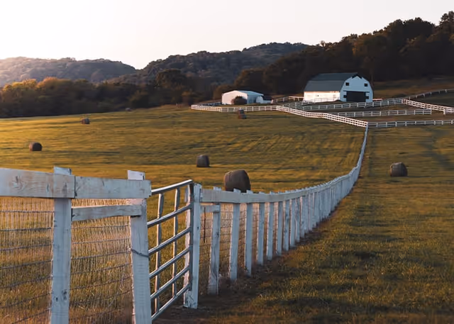 A white-fenced pasture with scattered hay bales leading toward a white barn and wooded hills in the background.