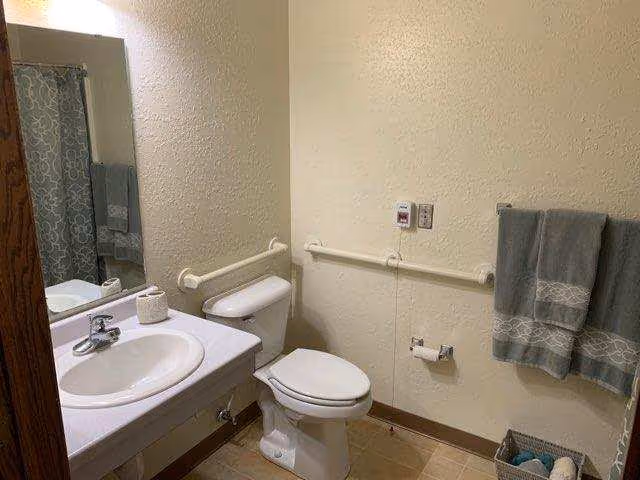 A bathroom with a white sink and countertop, a toilet with grab bars on the walls beside it, a towel rack with two gray towels, a basket with rolled towels on the floor, and a shower curtain with a gray and white pattern visible in the mirror reflection.