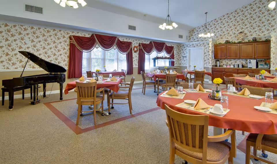 A dining room with multiple round tables covered with red tablecloths, each set with plates, napkins, cups, and glasses. Wooden chairs surround the tables. The room has floral wallpaper, large windows with red curtains, and a black grand piano in the corner. There are also wooden cabinets and a counter with various items on it along one wall.