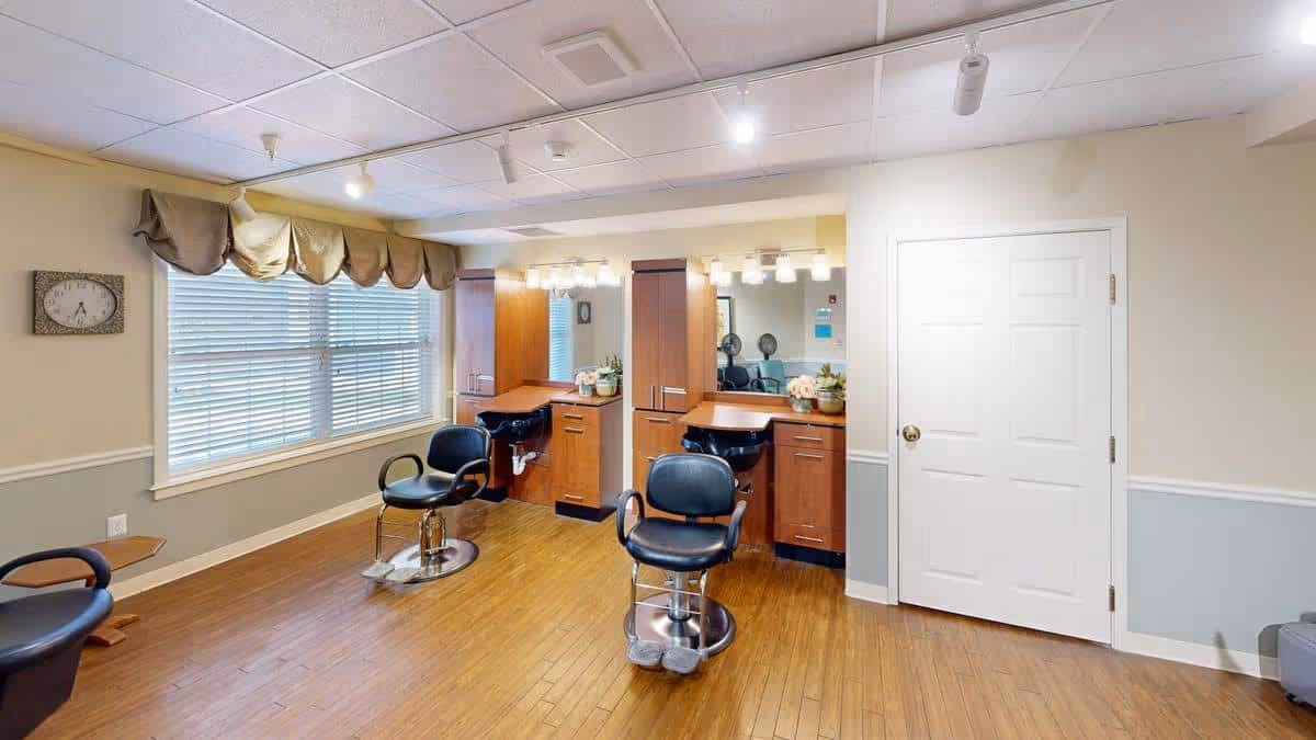 Interior view of a salon area in Pegasus Landing of Overland Park featuring two black salon chairs in front of wooden styling stations with mirrors and bright overhead lighting. The room has wood flooring, a large window with blinds and valance, and a white door on the right side.