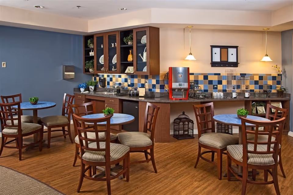 A cozy dining area with several round tables and wooden chairs with striped cushions. The back wall features a countertop with a coffee machine, cups, and decorative items. Above the counter are cabinets with glass doors displaying plates and plants. The walls are painted blue and beige with a tiled backsplash in blue, beige, and yellow tones. The floor is wooden, and two pendant lights hang from the ceiling.
