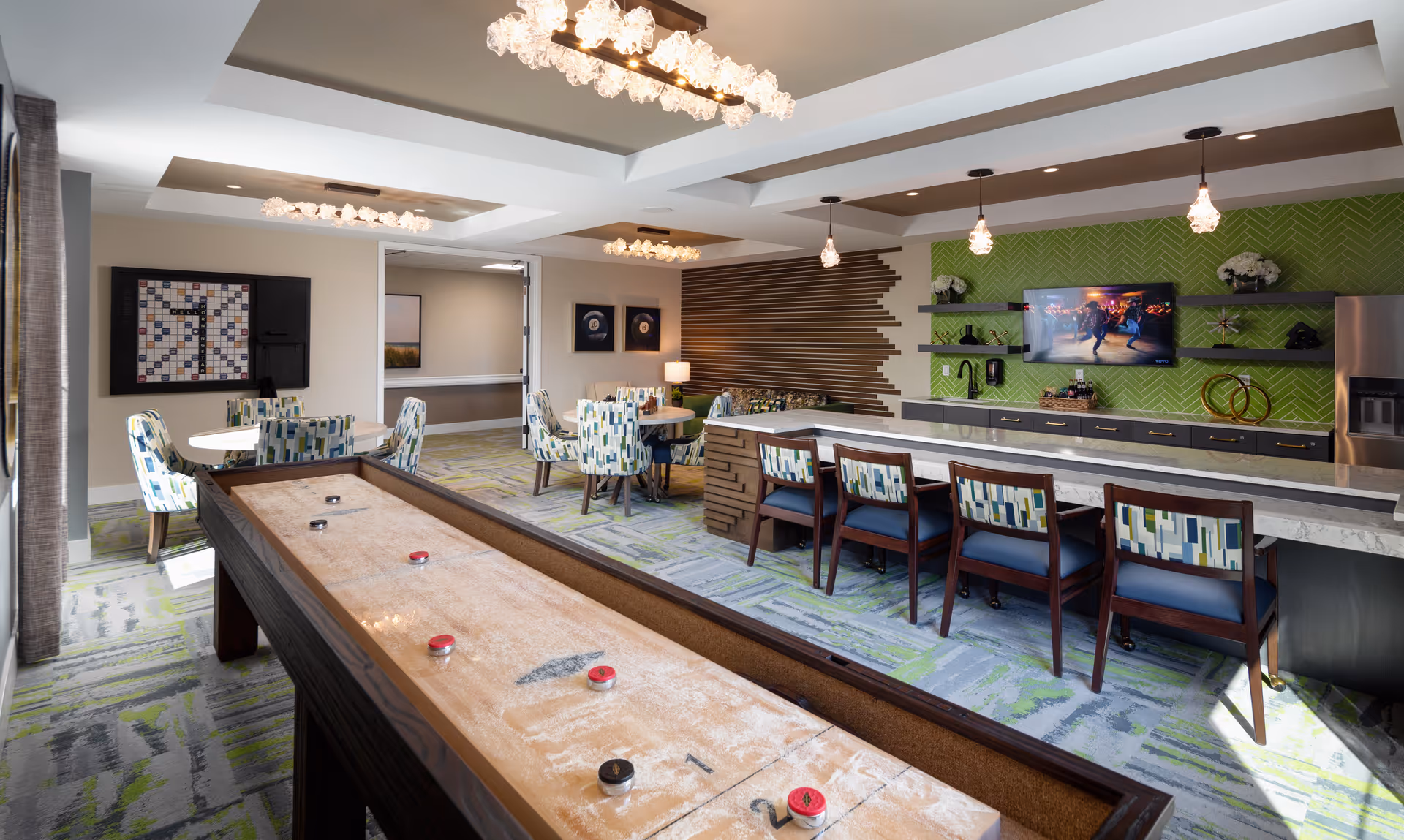 Bright, modern common room with a shuffleboard table in the foreground, a long bar with stools, seating tables, and a wall-mounted TV.