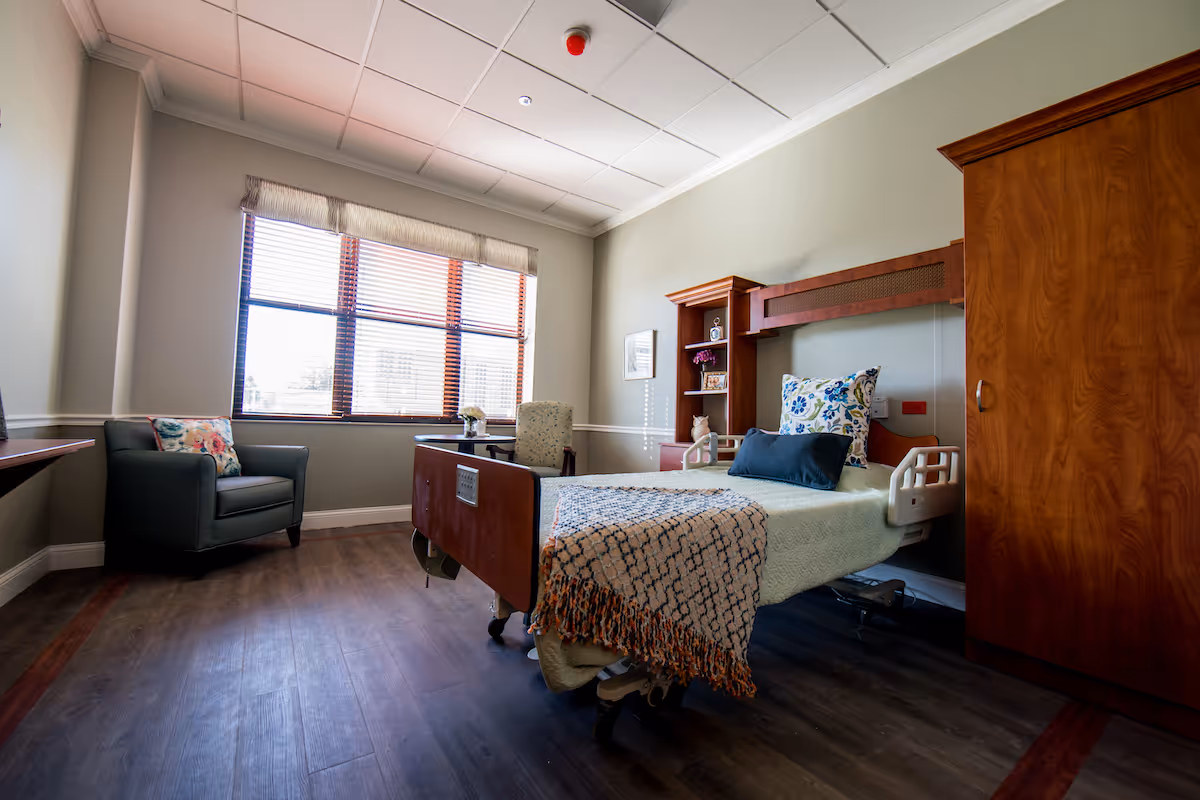 Well-lit resident bedroom with a hospital-style bed, armchair, window and wooden cabinetry.