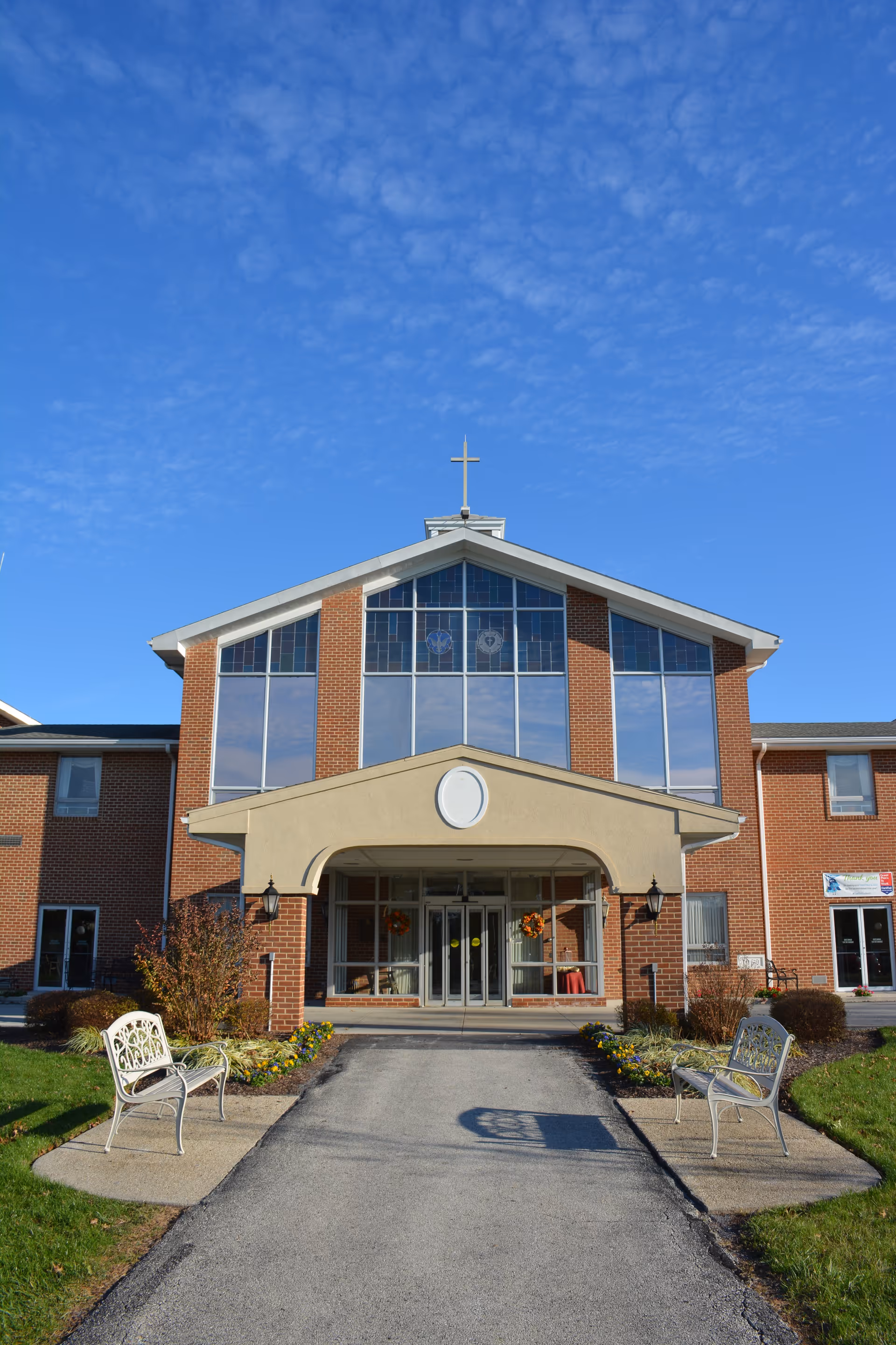 Front view of a brick senior living facility with a wide entrance, large store-front windows, and two white benches along a paved path under a bright blue sky.