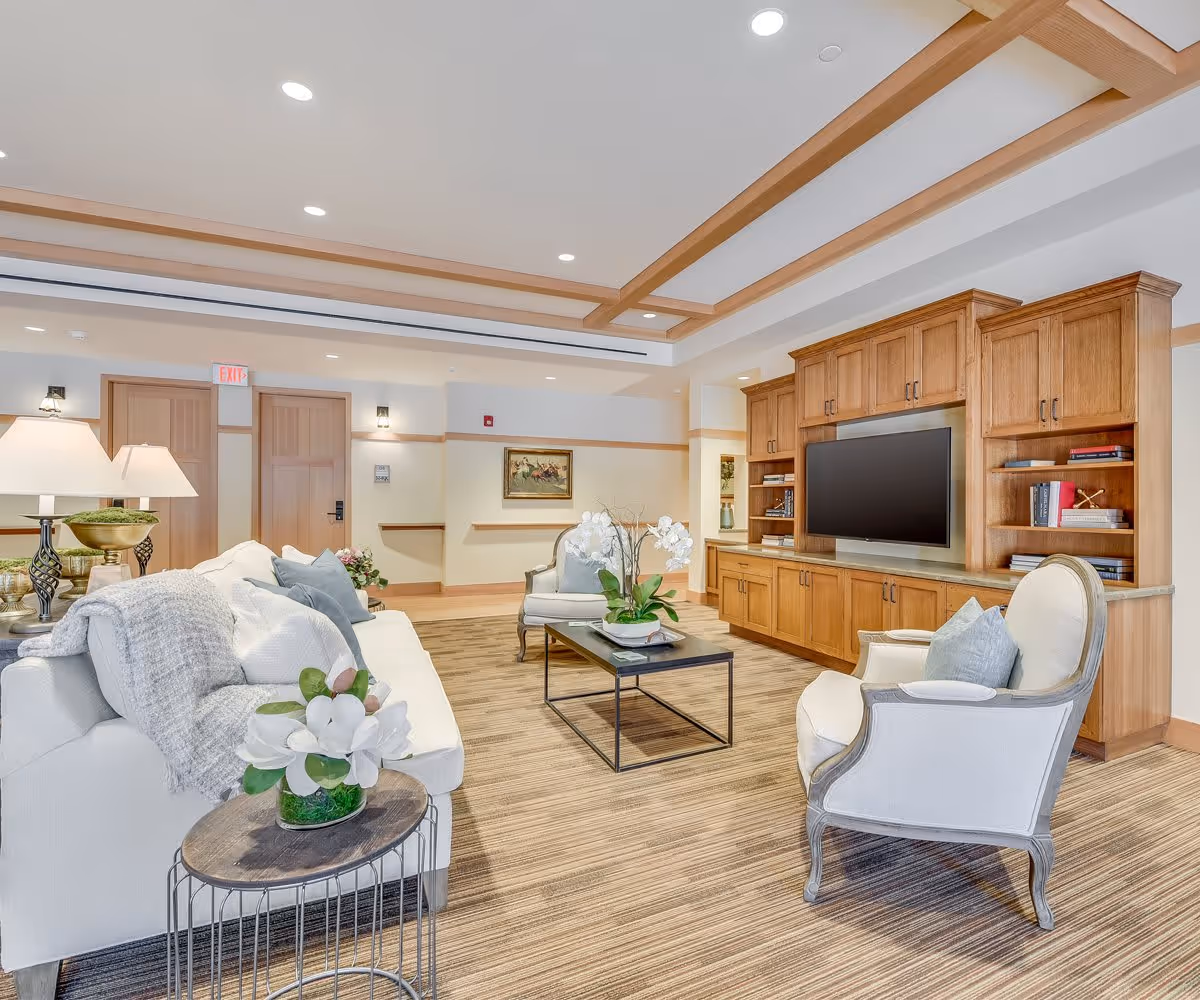 A bright and spacious living room in an assisted living facility featuring a white sofa with cushions and a throw blanket, two upholstered armchairs, a wooden coffee table with a white orchid centerpiece, and a large wooden entertainment center with a flat-screen TV. The room has a beige striped carpet, recessed ceiling lights, and wooden ceiling beams. There are two wooden doors and a framed painting on the far wall.