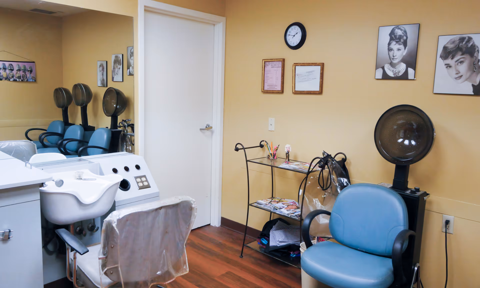 Interior of a hair salon room with blue salon chairs, hair dryers, a white sink, a small metal shelf with magazines and pencils, and framed black and white portraits on the beige walls.