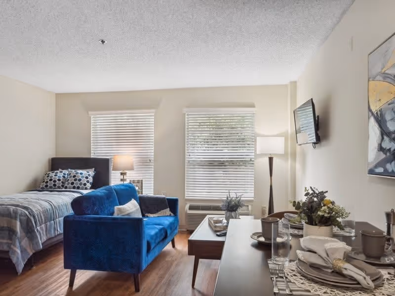 A cozy studio apartment with a bed, a blue loveseat, a coffee table, and a dining table set for four. Two windows with blinds allow natural light into the room. A floor lamp and a wall-mounted TV are visible, along with a piece of abstract artwork on the wall.