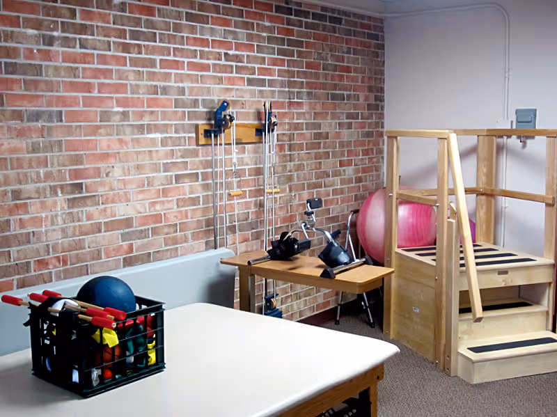 Rehabilitation room with a treatment table, a crate of exercise tools, resistance pulleys on a brick wall, an exercise bike, a stability ball, and wooden therapy stairs.