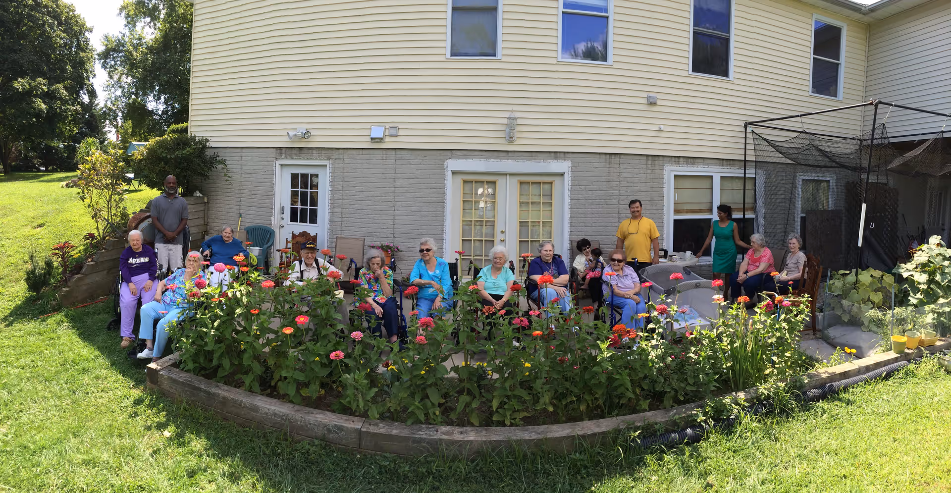A group of elderly people and two caregivers sitting and standing outside in a garden area in front of a building. There are colorful flowers in a raised garden bed in front of them, and the building has beige siding with several windows and doors.