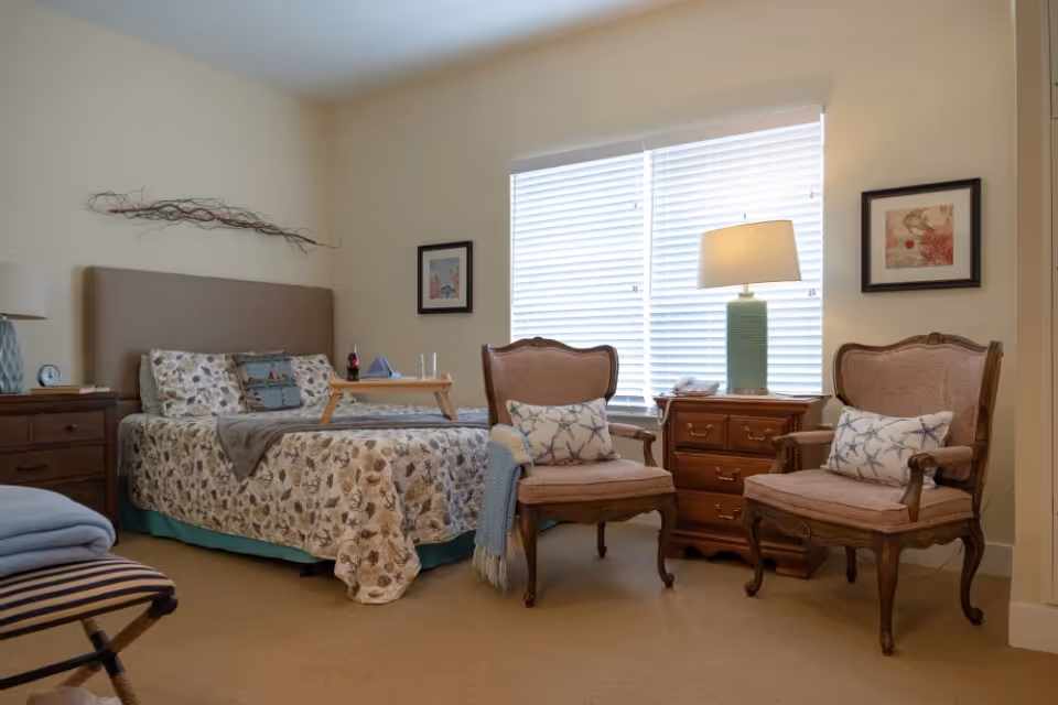 A cozy bedroom with a bed covered in a patterned quilt and pillows, a wooden nightstand with a lamp, two upholstered armchairs with decorative pillows, a wooden dresser with a lamp, and a window with closed blinds letting in natural light.