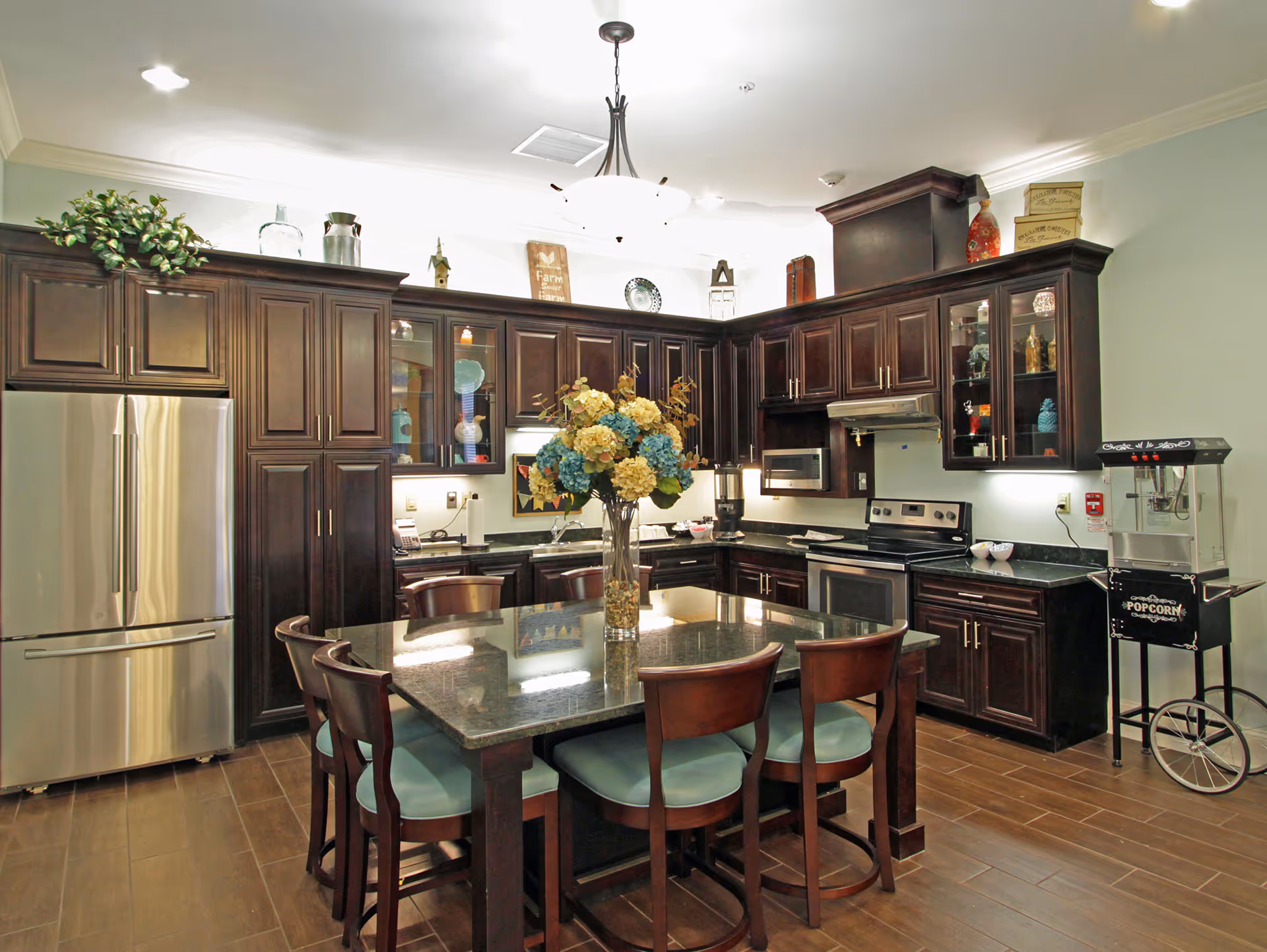 A well-lit kitchen with dark wooden cabinets, a stainless steel refrigerator, an electric stove, microwave, and a popcorn machine. In the center is a square dining table with six chairs and a vase of blue and yellow flowers on top. The floor is wooden, and decorative items are placed on top of the cabinets.