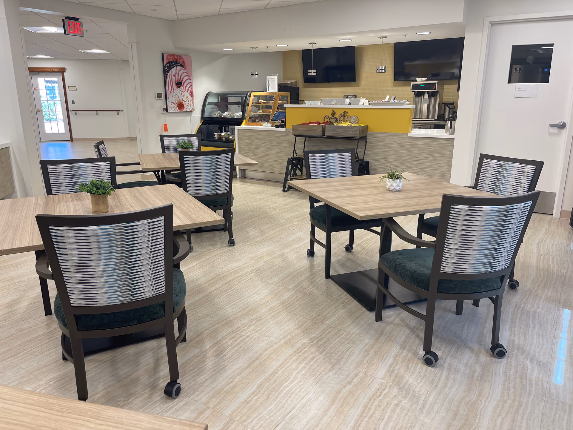 A clean and modern dining area with several square wooden tables and chairs with patterned backs and green cushions on wheels. Small potted plants are placed on the tables. In the background, there is a counter with a coffee machine, a display case with pastries, and two large flat-screen TVs mounted on a yellow-tiled wall. An exit sign and hallway are visible to the left.