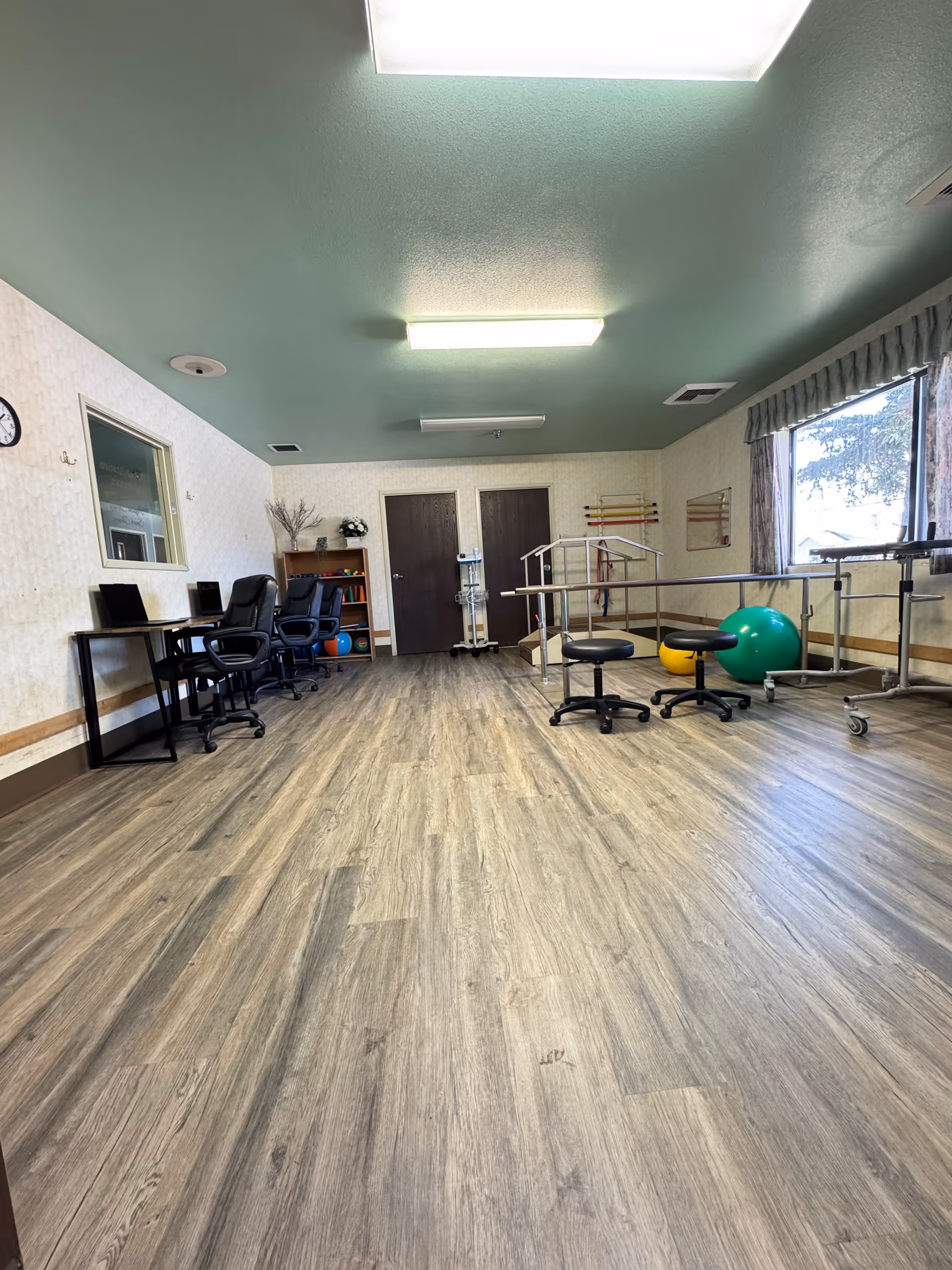 Spacious rehabilitation room with parallel bars, exercise balls, rolling stools and desks on wood-look flooring under a green ceiling.