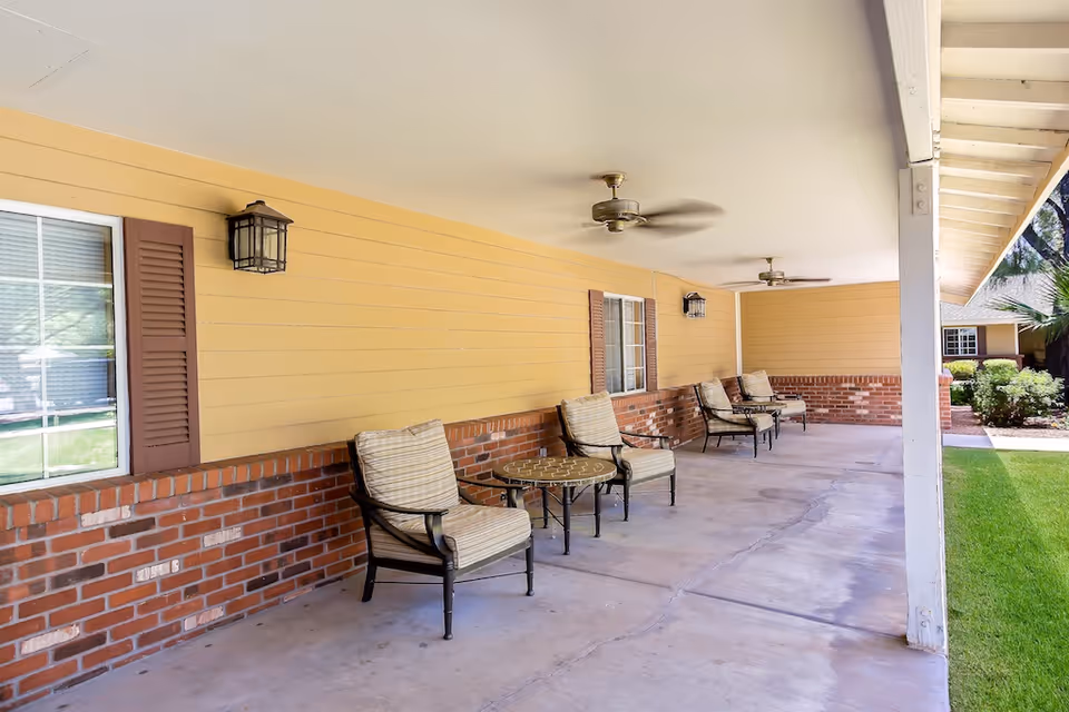 Covered outdoor porch with cushioned chairs and small tables along a yellow-sided building with ceiling fans.