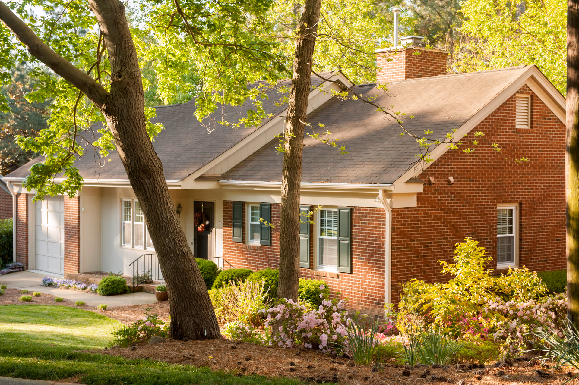 A single-story brick house with a sloped roof, white trim, and green shutters surrounded by trees and flowering bushes in a well-maintained garden.