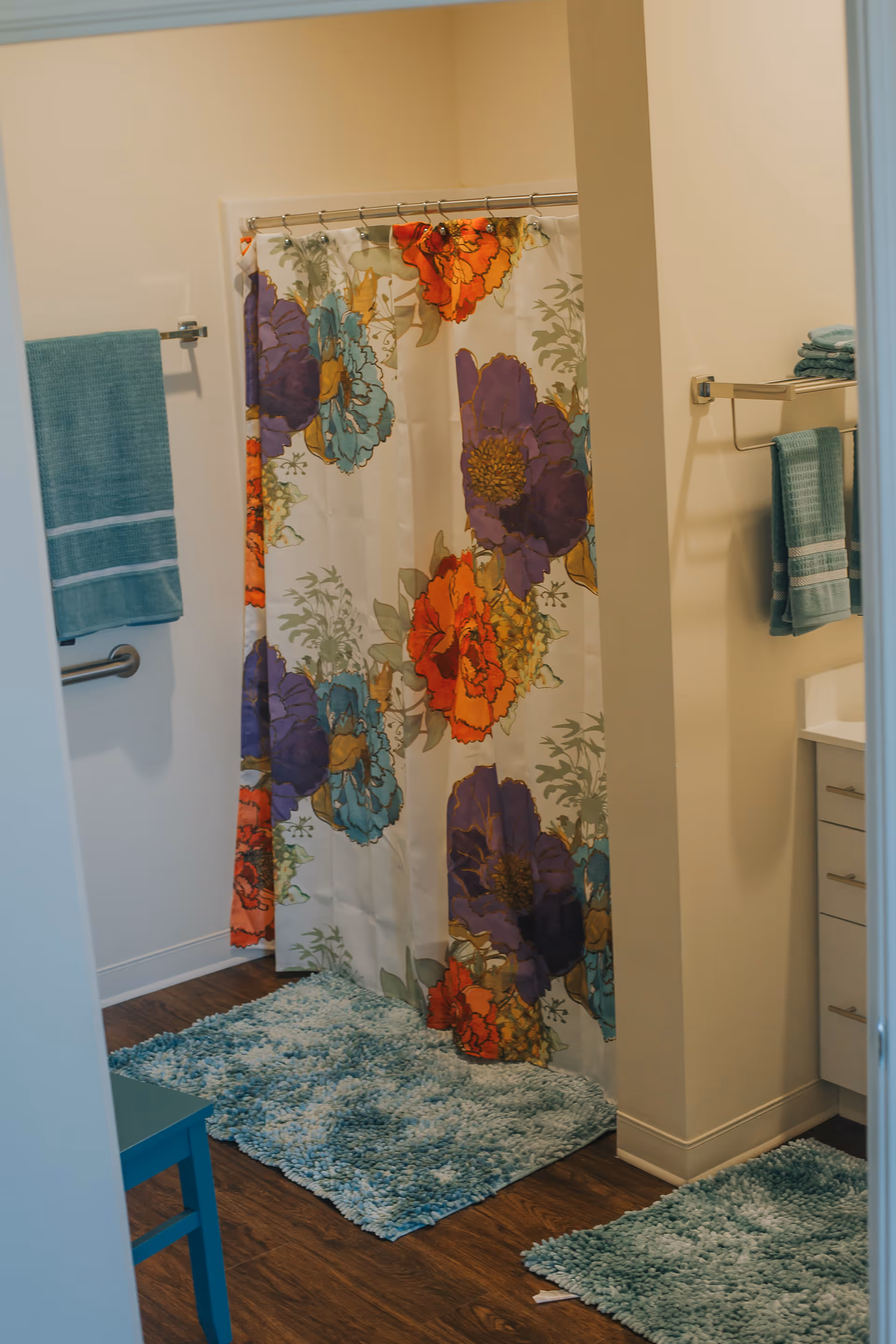 Bathroom with a colorful floral shower curtain, teal bath mats on a wooden floor, teal towels hanging on towel racks, and a white vanity with drawers.