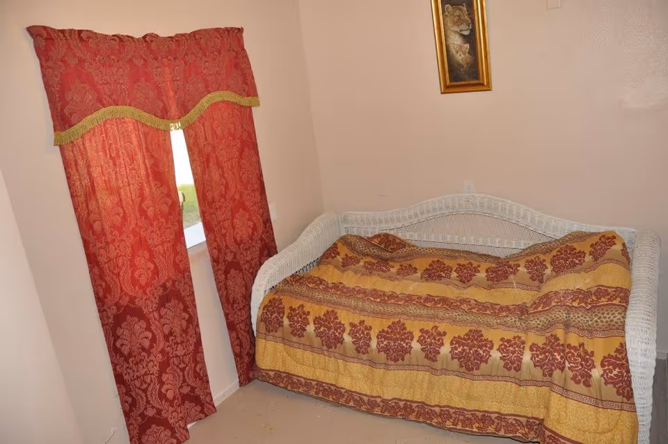 A small bedroom with a white wicker daybed covered by a yellow and red patterned blanket and a window with red drapes.