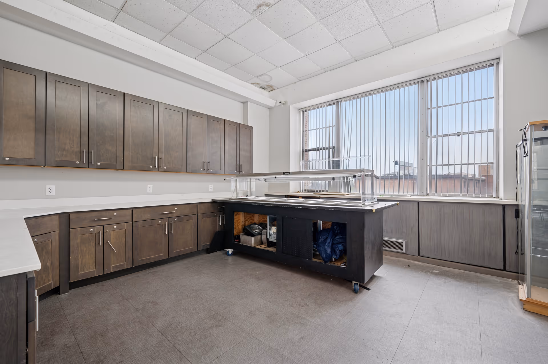 A spacious kitchen area with dark wooden cabinets along the walls and a central island with a glass sneeze guard. Large windows with vertical blinds allow natural light to fill the room. The floor is covered with a gray material, and the ceiling has white tiles.